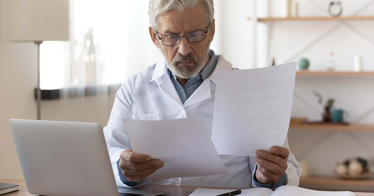 Healthcare worker with a white coat holding papers seated in front of laptop computer.
